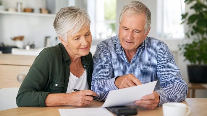 Senior couple sitting at a table reviewing and pointing at documents together in a bright, modern kitchen.